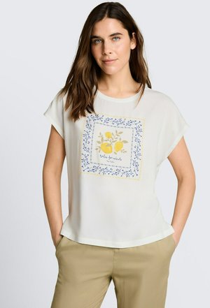 Kobieta ubrana w białą bluzkę z krótkim rękawem z kwadratowym wzorem cytryny i niebieskich kwiatów, w połączeniu z beżowymi spodniami, ręce w kieszeniach, neutralne tło.