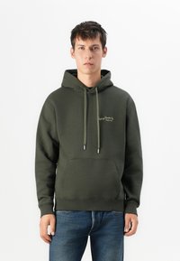 Young man standing, wearing a dark green hoodie with front pocket and blue jeans, against a plain white background.