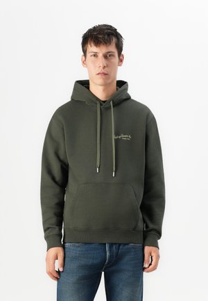 Young man standing, wearing a dark green hoodie with front pocket and blue jeans, against a plain white background.