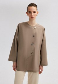 Beanie-style button-up shirt in beige, featuring large buttons, a round neckline, and wide sleeves, crafted from soft, smooth fabric.