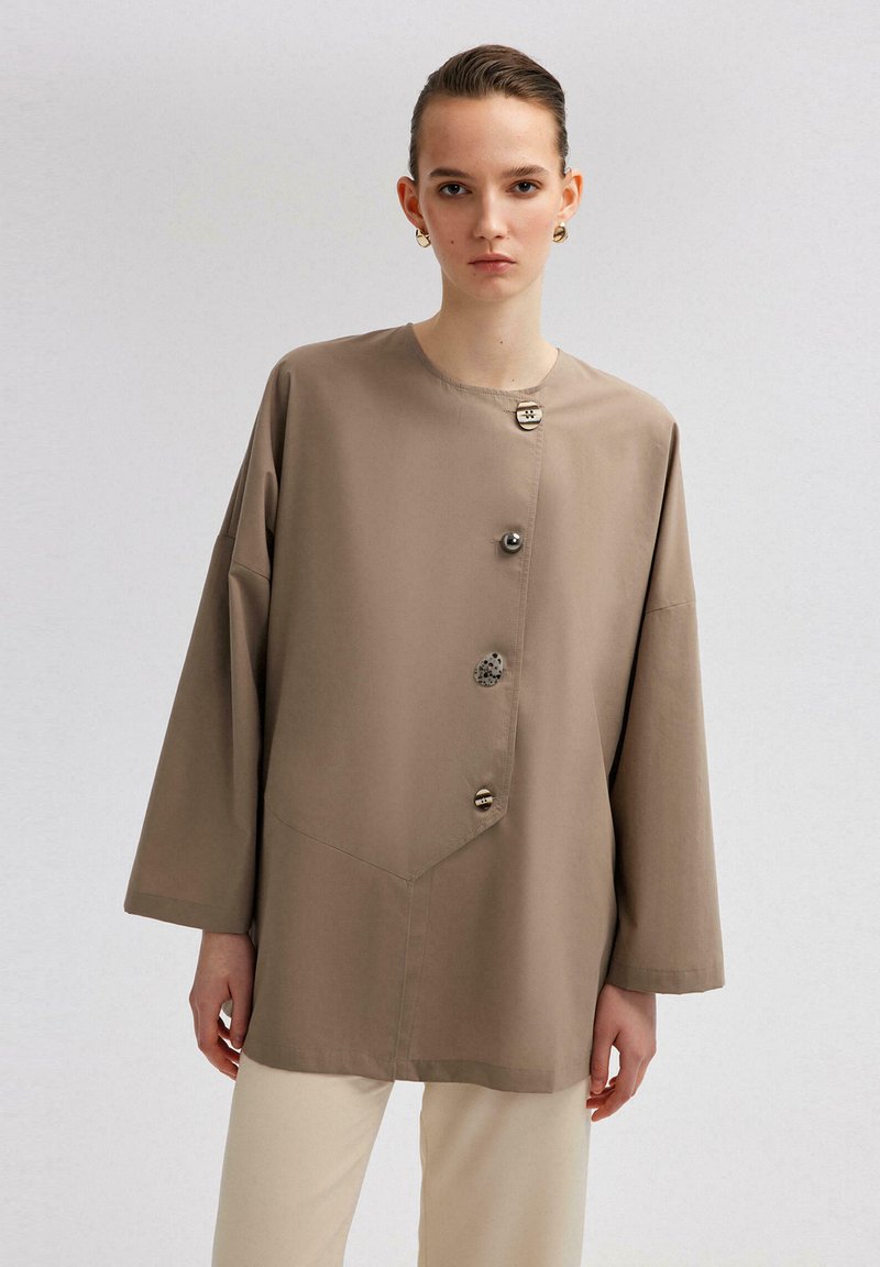 Beanie-style button-up shirt in beige, featuring large buttons, a round neckline, and wide sleeves, crafted from soft, smooth fabric.