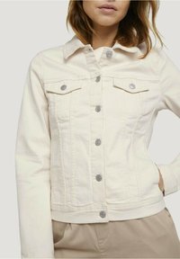 Giacca di denim beige chiaro con bottoni argentati, due tasche sul petto e colletto. Lunghezza corta, design aderente e dettagli di cucitura visibili.