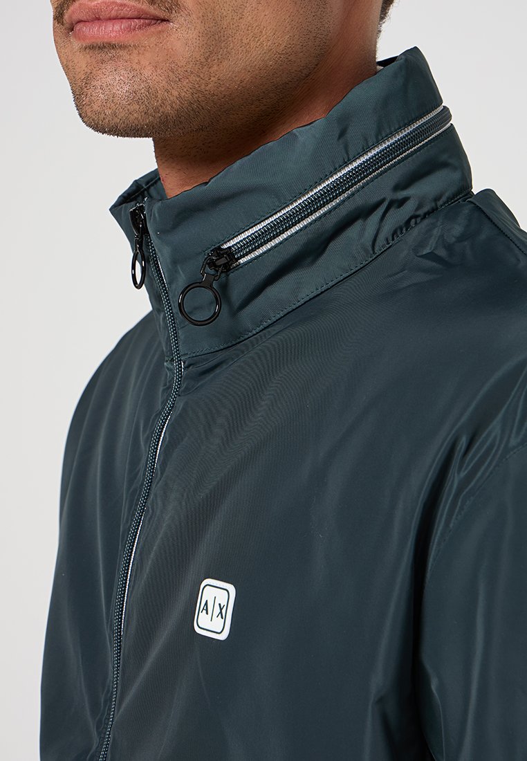 Giacca a vento verde scuro con colletto alto, taschino con zip sul petto e toppe con logo. Materiale lucido e leggero con texture minima.