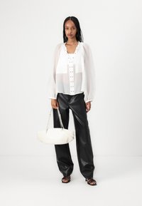 Blusa bianca trasparente con bottoni a pressione, abbinata a pantaloni in ecopelle nera a vita alta. Il modello tiene una borsa bianca a forma curvata.