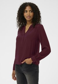 Bordeaux langærmet bluse med v-hals, knapdetaljer, blød tekstur og samlede ærmer, parret med mørke jeans.