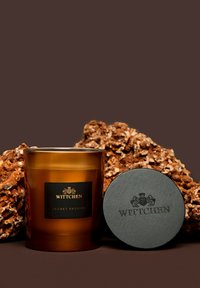 Candela in vetro ambra etichettata "WITTCHEN SECRET PASSION," con un coperchio nero con logo inciso; posizionata su rocce marroni texture.