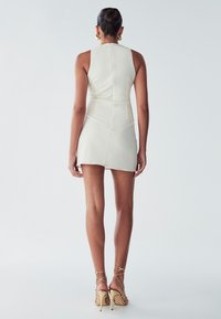 Robe mini blanche sans manches avec un tissu texturé, design ajusté et un col montant. Associée à des talons beiges à lanières.
