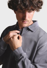 Šedý pulóver s golierom, s rebrovanými manžetami a malým logom slona na hrudi. Materiál sa zdá byť mäkký a s textúrou.
