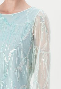 Top azzurro chiaro trasparente con dettagli intricati in frange di paillettes. Presenta un collo rotondo e maniche lunghe e traslucide.