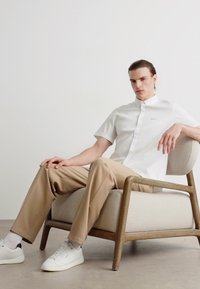 Camicia bianca a maniche corte con un logo sottile, pantaloni beige e sneakers bianche. Il modello è seduto su una sedia in legno chiaro.