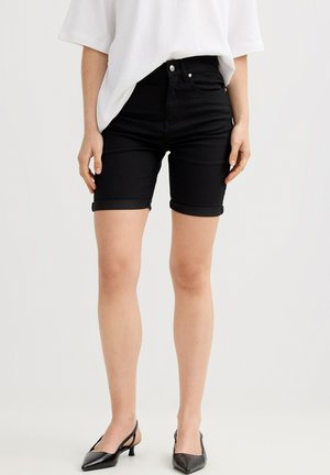 Persona con pantalones cortos de mezclilla negros remangados, camisa blanca holgada y zapatos slingback negros de tacón bajo y punta afilada, de pie frente a un fondo liso.