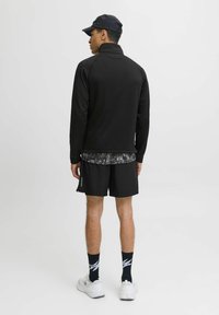 Sudadera negra de manga larga con cuello alto, llevada sobre una camisa de estampado, combinada con pantalones cortos negros, zapatillas blancas y una gorra azul marino.