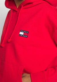 Czerwona bawełniana bluza z kapturem z kieszenią z przodu, sznurkiem do ściągania i prostokątną łatą z logo "TOMMY JEANS" w kolorze białym i granatowym.