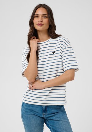 Femme aux longs cheveux foncés portant un t-shirt rayé blanc et bleu avec un cœur noir et un jean bleu, posant avec une main près du visage.