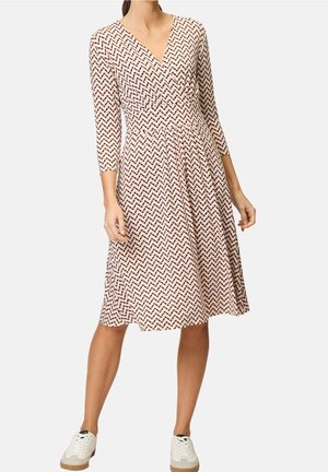 Frau trägt ein knielanges, langärmliges Kleid mit Muster und V-Ausschnitt, kombiniert mit weißen Turnschuhen, steht vor einem schlichten Hintergrund.