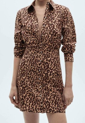 Frau trägt ein braun-schwarzes, leopardenmusterndes Hemdblusenkleid mit langärmeligen Ärmeln, die bis zu den Ellbogen hochgekrempelt sind, und steht vor einem schlichten Hintergrund.