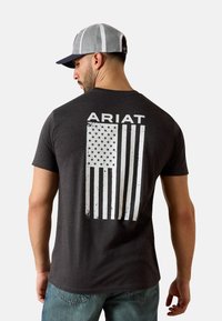 Camiseta gris oscuro con texto blanco "ARIAT" y un gráfico de la bandera americana desgastada en la parte trasera. La tela parece suave con un ajuste clásico.