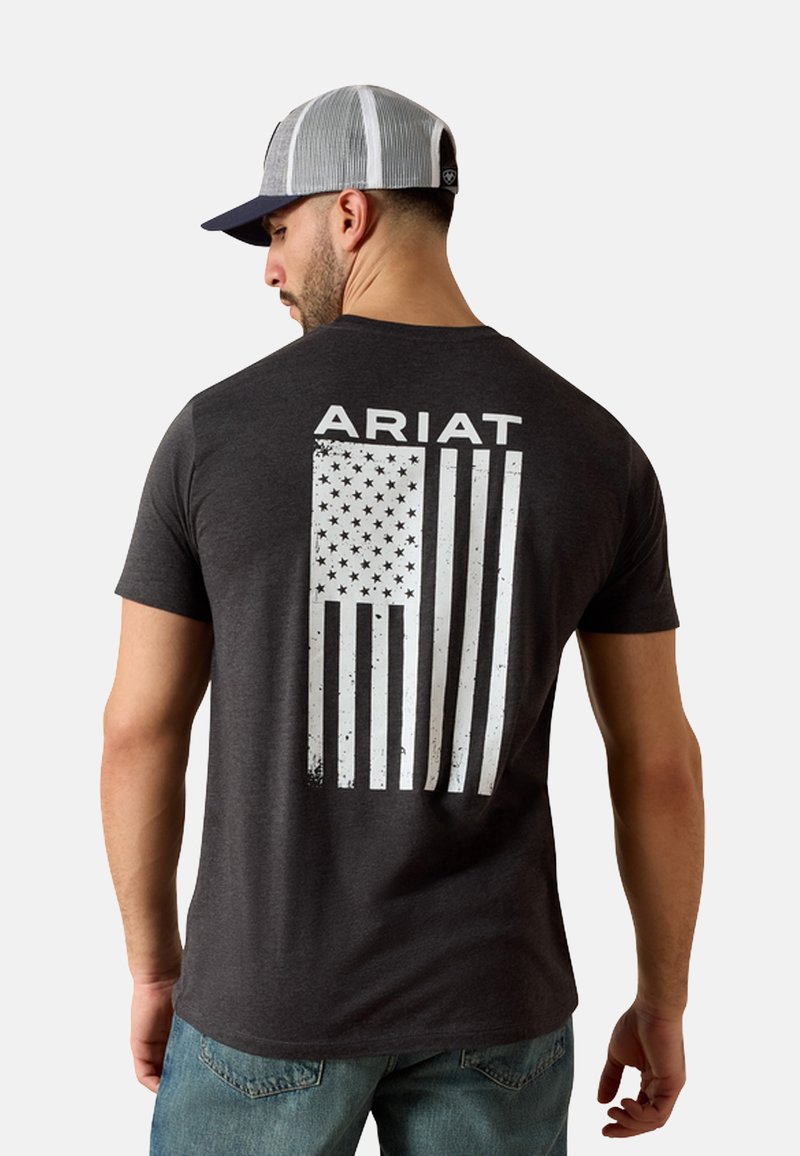 Camiseta gris oscuro con texto blanco "ARIAT" y un gráfico de la bandera americana desgastada en la parte trasera. La tela parece suave con un ajuste clásico.
