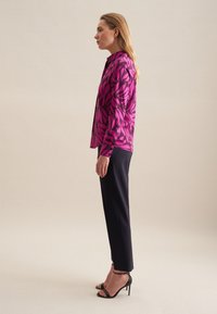 Intensywnie różowa koszula z długim rękawem w czarny wzór liści, zestawiona z dopasowanymi granatowymi spodniami i czarnymi szpilkami, widok z boku.