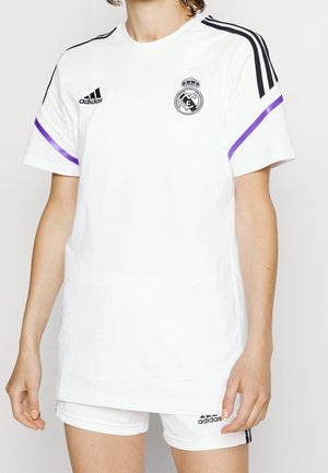 Person iført hvid Adidas Real Madrid sportstrøje med sorte logoer, lilla ærmestriber og matchende hvide shorts.