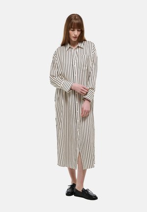 Femme ajustant le poignet d'une robe chemise longue crème à rayures verticales, avec col, poche avant et chaussures noires à lacets sur fond blanc.