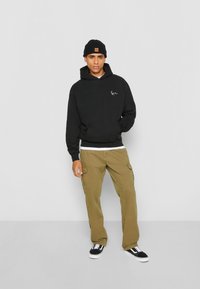 Hoodie noir avec poche avant et logo brodé, associé à un pantalon cargo olive. Le modèle porte un bonnet noir et des baskets noires avec des semelles blanches.