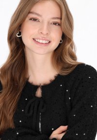 Czarny dziergany kardigan o puszystej fakturze, z dekoltem w kształcie łezki, z wiązaniem z przodu i rozproszonymi akcentami w postaci cekinów.