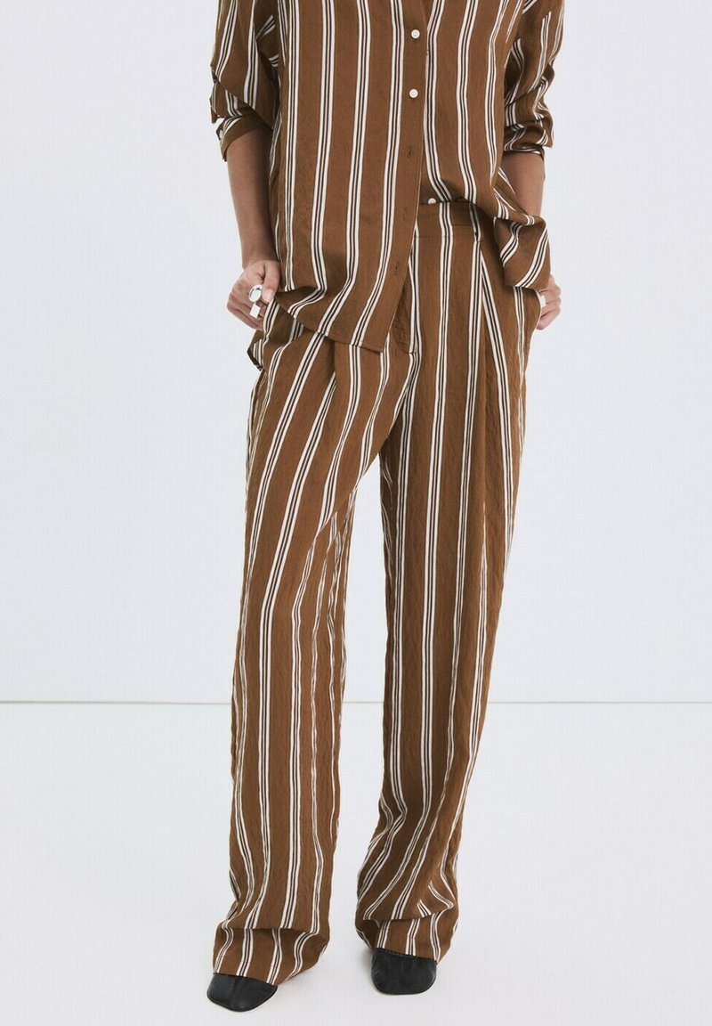 Pantalones marrones sueltos y camisa de manga larga a juego con rayas verticales blancas, combinados con zapatos negros puntiagudos.