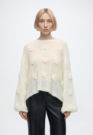 Ung person med kort blondt hår iført en hvid, tekstureret, oversized sweater og sorte læderbukser, stående mod en ensfarvet baggrund.