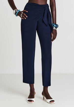 Pantaloni avvolgenti di un blu scuro indossati da una persona con braccialetti in argento e blu a strati e sandali con tacco aperti bianchi.
