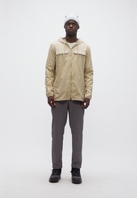 Άνδρας που στέκεται και κοιτάζει μπροστά, φοράει μπεζ μπουφάν με κουκούλα, γκρι παντελόνια, μαύρα παπούτσια και ανοιχτό γκρι σκουφί, μπροστά σε λευκό φόντο.