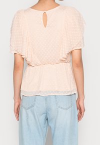 Femme portant un blouse rose clair à manches courtes avec un dos en trou de serrure et des manches volantes, assortie à un jean en denim bleu clair.