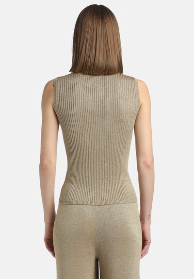 Femeie cu părul drept, până la umeri, purtând un top din material ribbed, aurie, fără mâneci și pantaloni aurii asortați, întoarsă cu fața spre un fundal alb.