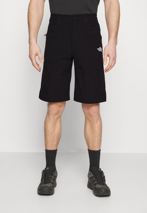 Homme portant un short cargo noir avec le logo The North Face, des chaussures de randonnée noires, des chaussettes noires et un t-shirt noir, debout devant un fond uni.