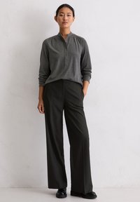 Langärmliges graues Henley-Shirt mit Knöpfen, kombiniert mit hoch taillierten, weit geschnittenen schwarzen Hosen und schwarzen Schuhen, vor einem hellen Hintergrund.