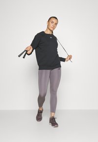 Sudadera negra de mangas largas y puños acanalados, combinada con leggings de color púrpura claro. Zapatillas deportivas oscuras, sosteniendo una cuerda para saltar.