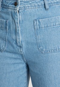 Světlé modré denimové džíny s předními kapsami, zapínáním na zip a jemnou strukturou. Materiál má tkaný vzor a klasický střih.
