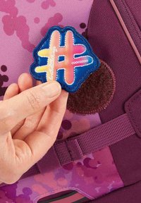 Coocazoo backpack in purple with a camouflage pattern. The hand is placing a colorful, stitched hashtag patch on a fuzzy area.
