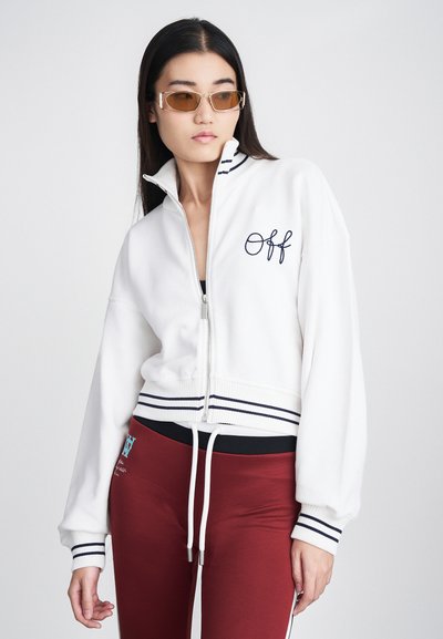 Sweatshirt blanc à zip avec "Off" brodé en bleu marine, doté de poignets et d'une taille rayés bleu marine. Porté avec un pantalon rouge, mettant en valeur un look sportif.