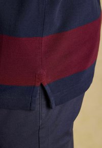 Svītrota polo krekla dizains tumši zilā un bordo krāsā, ar sānu venti un teksturētu audumu. Iezīmējas ar izteiksmīgu krāsu atdalījumu un šūšanas detaļām.