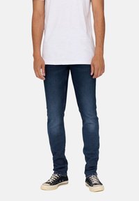 Tmavě modré slim-fit džíny vyrobené z denimu, s klasickým designem pěti kapes a nošené s černými plátěnými teniskami.