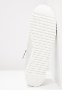 Semelle de basket blanche avec motif de bande de roulement rainurée horizontale, inclinée contre un mur d’angle blanc.