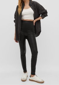 Leggings de imitación cuero negros de cintura alta combinados con una camiseta corta blanca y una camisa de mezclilla negra holgada. Zapatillas blancas con acentos beige.