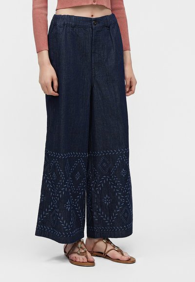 Femme portant un jean bleu foncé à jambes larges avec des motifs géométriques brodés près de l'ourlet, associé à des sandales marron et un pull rose court.