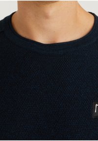 Granatowy sweter z teksturowanego dzianiny z okrągłym dekoltem i małą czarną metką marki z przodu. Gładki materiał z subtelnym wzorem.