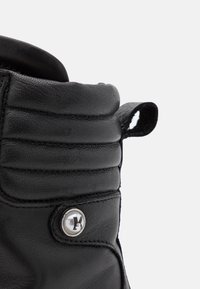 Bota de cuero negra con textura acanalada cerca de la parte superior, detalle de botón plateado con un logo y una pequeña tira de agarre en el lateral.