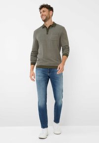 Olivgrüner strukturierter Polo-Pullover mit einer Brusttasche, kombiniert mit blauen schmal geschnittenen Jeans und weißen Sneakers. Schlichtes Design und entspannte Passform.