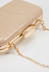 Clutch rectangular en suave tela beige con una textura brillante. Presenta herrajes dorados y una correa de cadena desmontable. Cierre con un broche redondo.
