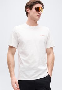 Νεαρός άνδρας που φοράει λευκό t-shirt Columbia και αντανακλαστικά πορτοκαλί αθλητικά γυαλιά ηλίου, στέκεται με το ένα χέρι στην τσέπη.