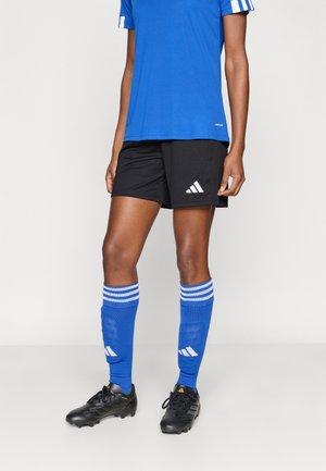 Persona con calzini da calcio blu, pantaloncini neri e maglia blu, in piedi con scarpe da calcio nere su uno sfondo semplice.